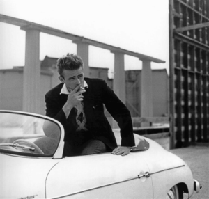 “James Dean driving his 1955 Porsche Super Speedster, a sleek silver sports car, embodying his passion for speed and freedom.”