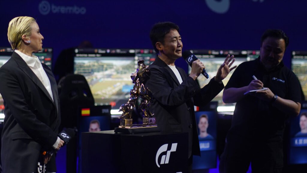 Kazunori Yamauchi discussing Gran Turismo and PlayStation’s legacy with a backdrop of sim racing visuals and cars