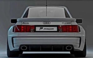 Classic Audi Quattro in white rally trim with flared fenders, parked on gravel with mountains in the background