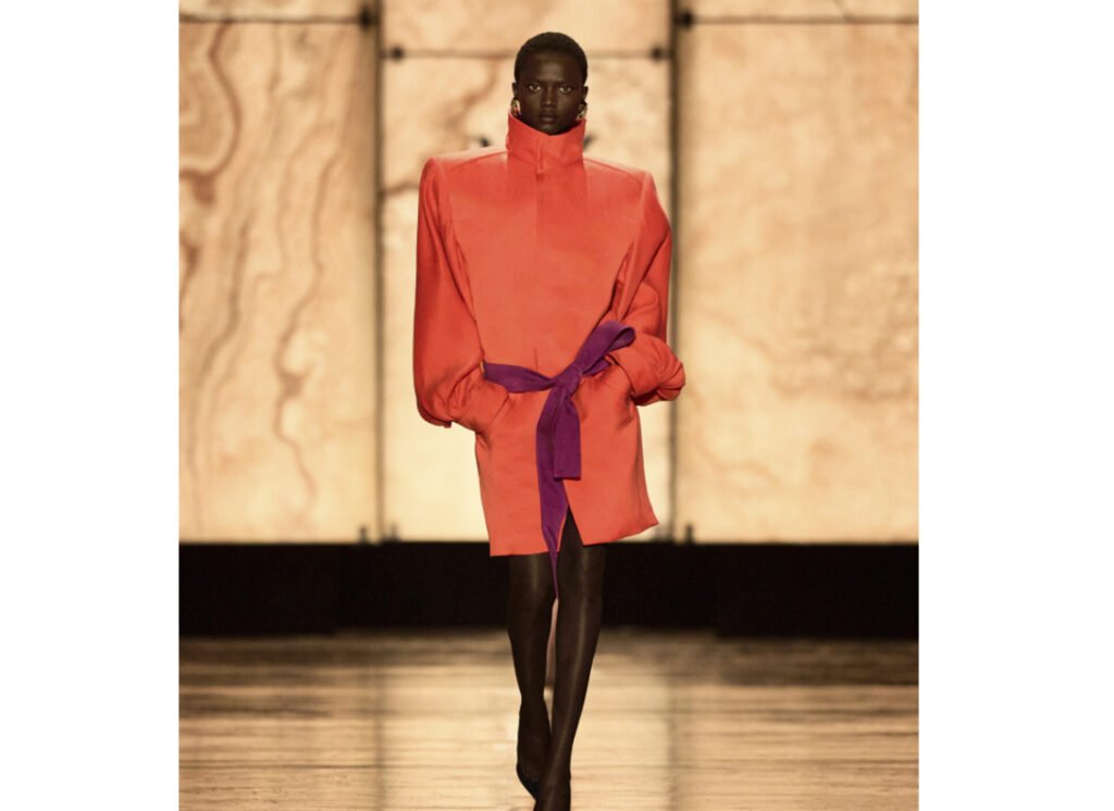 Model walks the Saint Laurent FW25 runway wearing a high-collared coat dress with soft lighting and a rock crystal backdrop