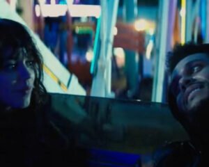 The Weeknd and Jenna Ortega in a vintage car at night in the “Drive” music video, surrounded by neon lights