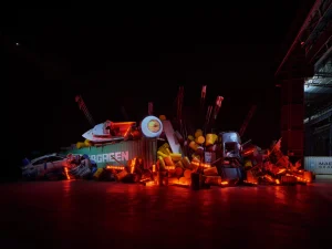 Large-scale art installation of piled boats, cars, barrels, and debris illuminated in red light, part of Yukinori Yanagi’s "Project Godzilla 2025