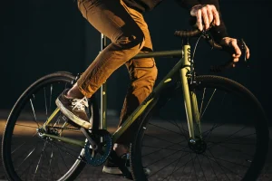 Close-up of a rider on a matte olive State Bicycle Co. 4130 fixed-gear bike, showing the frame details, drop handlebars, and urban riding posture in warm evening light