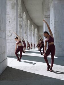 A line of women athletes dressed in matching burgundy NikeSKIMS sports bras and leggings performing synchronized side stretches beneath tall concrete arches, blending strength and elegance in a modern architectural setting.
