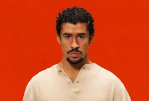 Bad Bunny photographed against a solid red background, wearing a cream-colored buttoned shirt, with a serious expression and short curly hair.