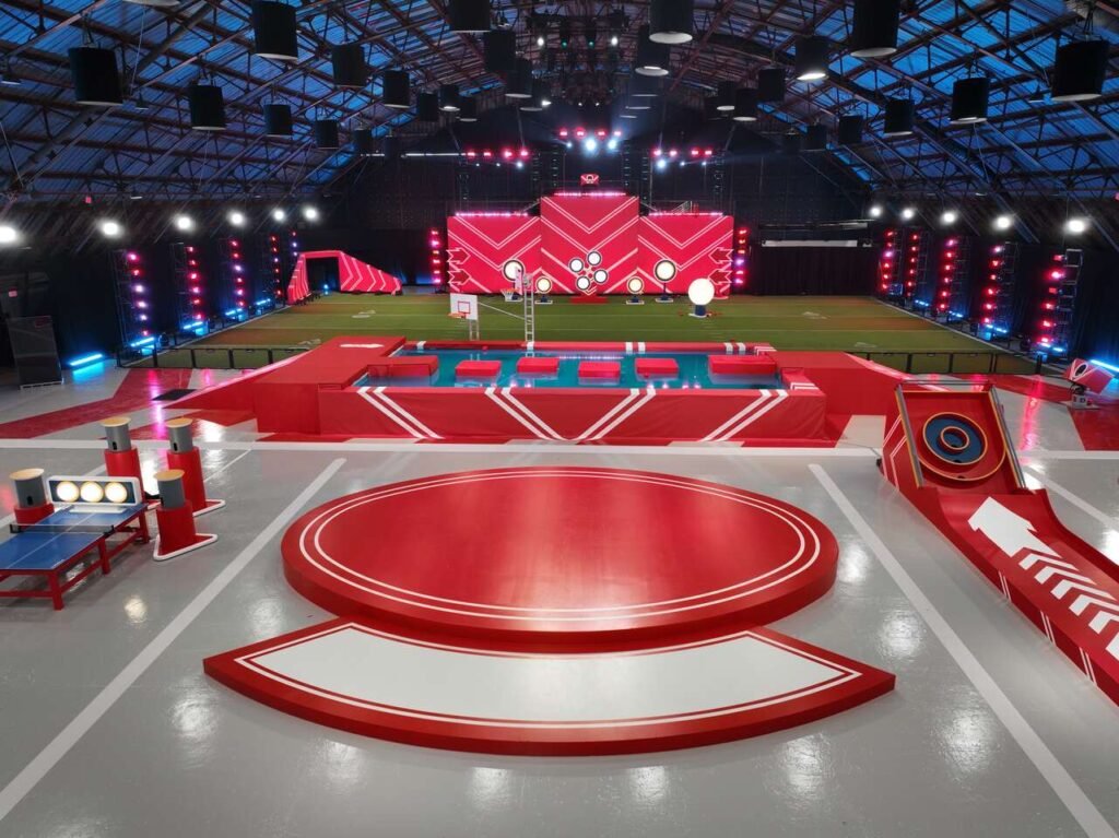 Play it Forward game show set featuring bright red obstacle courses, splash challenges, and stage lighting inside a large arena.