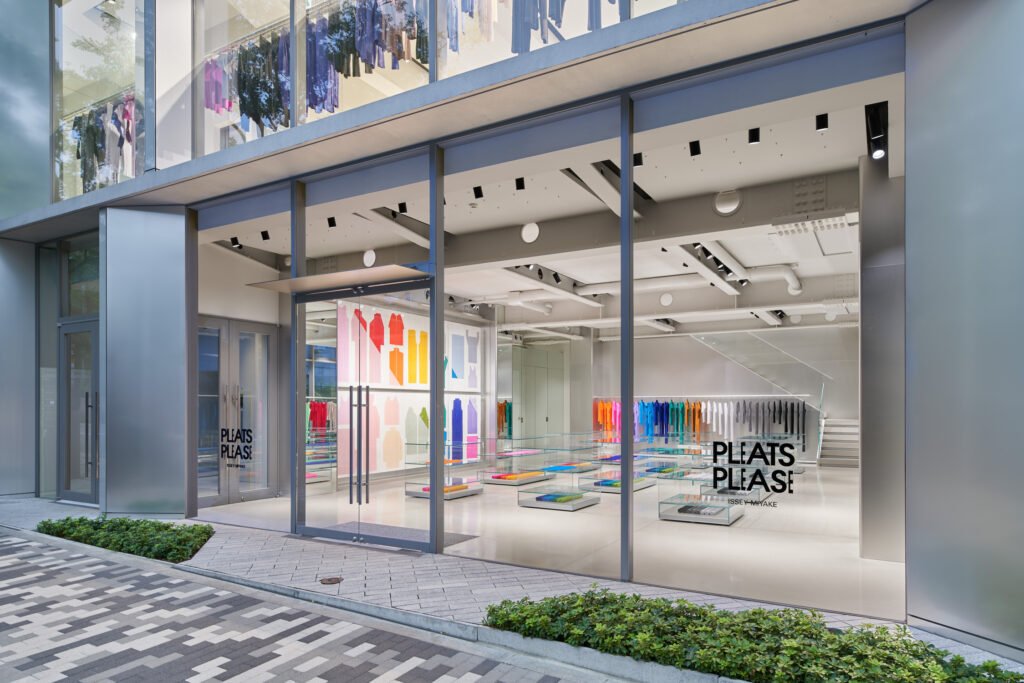 Exterior view of the new PLEATS PLEASE ISSEY MIYAKE flagship store in Aoyama, Tokyo, designed by Tokujin Yoshioka. The minimalist glass façade reveals a luminous interior with vibrant pleated garments arranged by color, reflecting the brand’s signature fusion of light, motion, and Japanese design.