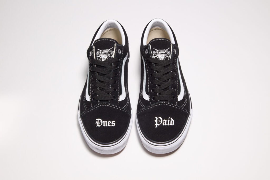 top-down view of the Travis Barker x Vans Old Skool “Dues Paid” sneakers showing black uppers with white side stripes, “Dues” and “Paid” embroidered across the toe boxes in gothic lettering, and drum set graphic printed on the tongue tags