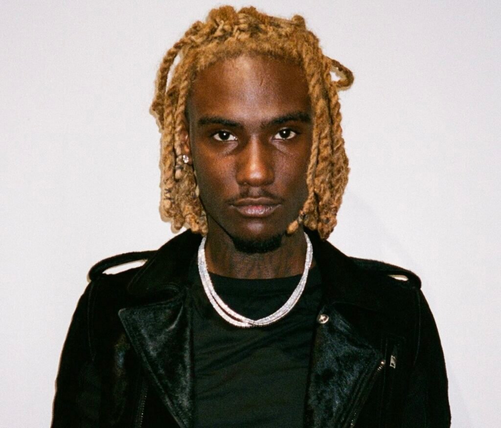 OsamaSon wearing a black jacket and layered silver chains, standing against a plain white background.