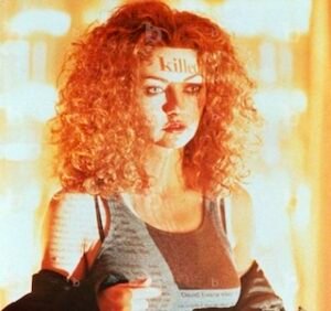 Scene from Urban Legend (1998) featuring a female character with curly red hair standing in front of glowing light, with the word ‘killed’ projected across her forehead.