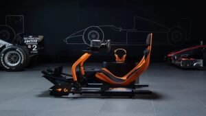 Side profile of the PLAYSEAT® Formula Instinct McLaren Racing Edition simulator, featuring an aerodynamic papaya-orange frame with integrated racing seat, steering wheel mount, and pedal base, set against a dark backdrop with Formula 1 car outlines and racing tires.