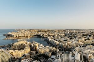 A panoramic aerial view of St Julian’s and Sliema on the island of Malta, showing the curving bay surrounded by golden limestone buildings, marinas, and boats under a clear blue Mediterranean sky at sunset.