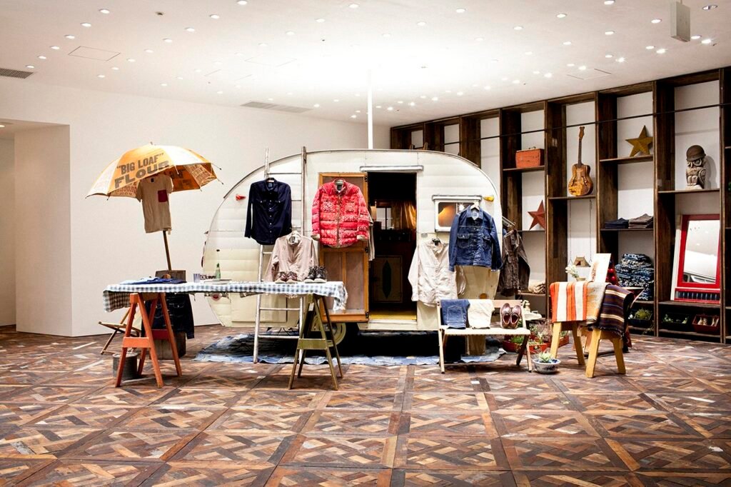 Interior view of an Indigo Camping Trailer pop-up installation featuring a vintage white camper surrounded by Visvim garments. A checkered table displays shirts and jackets dyed with indigo and mud, while shelves in the background hold guitars, textiles, and rustic décor, reflecting the brand’s fusion of craftsmanship, travel, and natural materials.