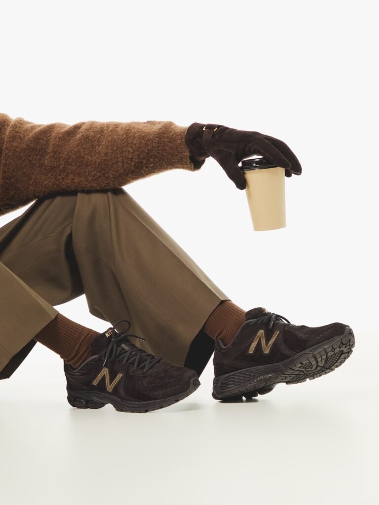 Model wearing the Kith x New Balance 860v2 sneakers in rich brown suede, styled with tailored trousers, brown knitwear, and gloves while holding a coffee cup — showcasing refined street-haute aesthetics.