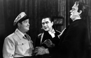 A black-and-white still from the 1948 film Abbott and Costello Meet Frankenstein. The scene features three characters: a smiling man in a uniform cap facing a vampire in a dark cape and the towering Frankenstein Monster. The vampire appears to be speaking to the uniformed man while the Monster looms nearby, creating a humorous yet classic blend of comedy and horror characteristic of the film.
