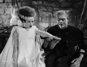 A black-and-white still from the 1935 film Bride of Frankenstein, depicting the Monster and his newly created Bride in the laboratory. The Bride, wrapped in a flowing white gown with streaked hair standing tall, recoils in surprise as the Monster gently reaches out his hand toward hers. The scene is illuminated by eerie lab equipment in the background, capturing both the gothic atmosphere and tragic emotion of the moment.