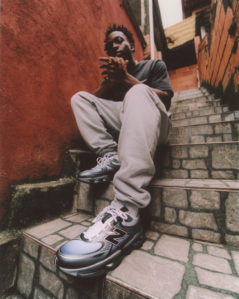 A person sits on stone steps wearing the New Balance ABZORB 2000 sneakers in the “Pearl Grey with Black” colourway. The metallic grey and black shoes stand out against the rustic red and brick alley backdrop, highlighting their sleek, performance-inspired design.