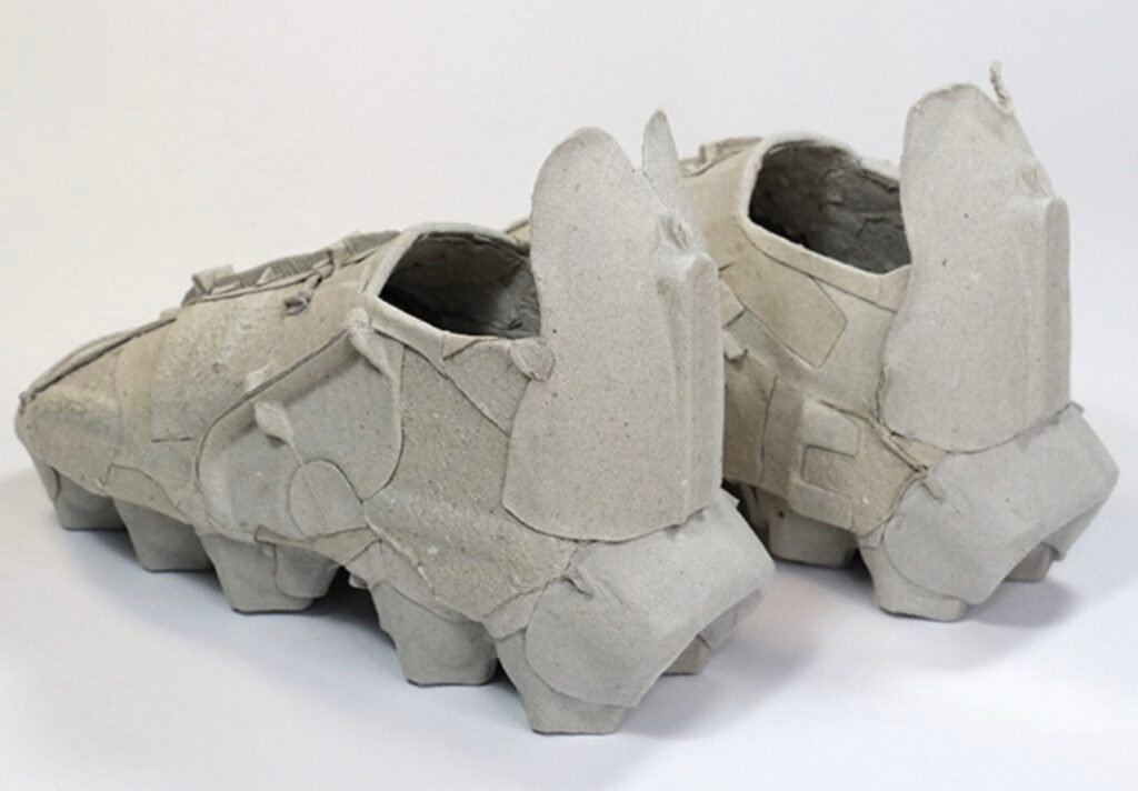 Rear view of Vadim Kibardin’s Egg Box Sneakers, made from molded recycled cardboard. The shoes feature angular, overlapping panels that resemble fragments of egg cartons, with raised heel structures and textured seams emphasizing their handmade construction. Displayed on a neutral white background, the image highlights the sculptural quality and experimental use of waste material as a design medium.