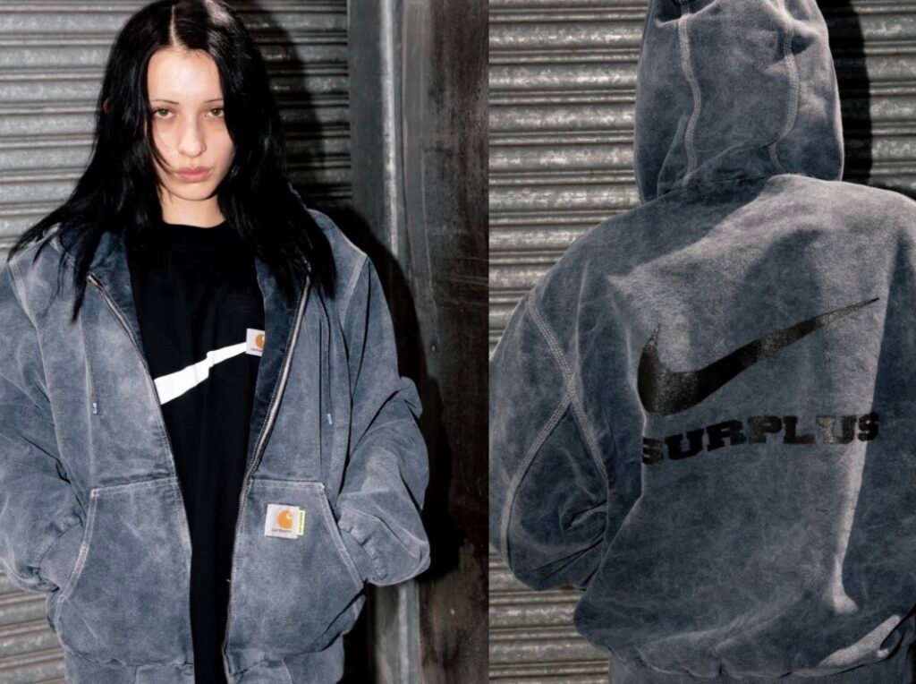 Model wearing the Carhartt WIP × Nike SURPLUS® 2025 washed grey canvas jacket layered over a black tee, showing the front Carhartt label and back view with oversized black Nike Swoosh and ‘SURPLUS’ lettering printed across the hooded jacket.