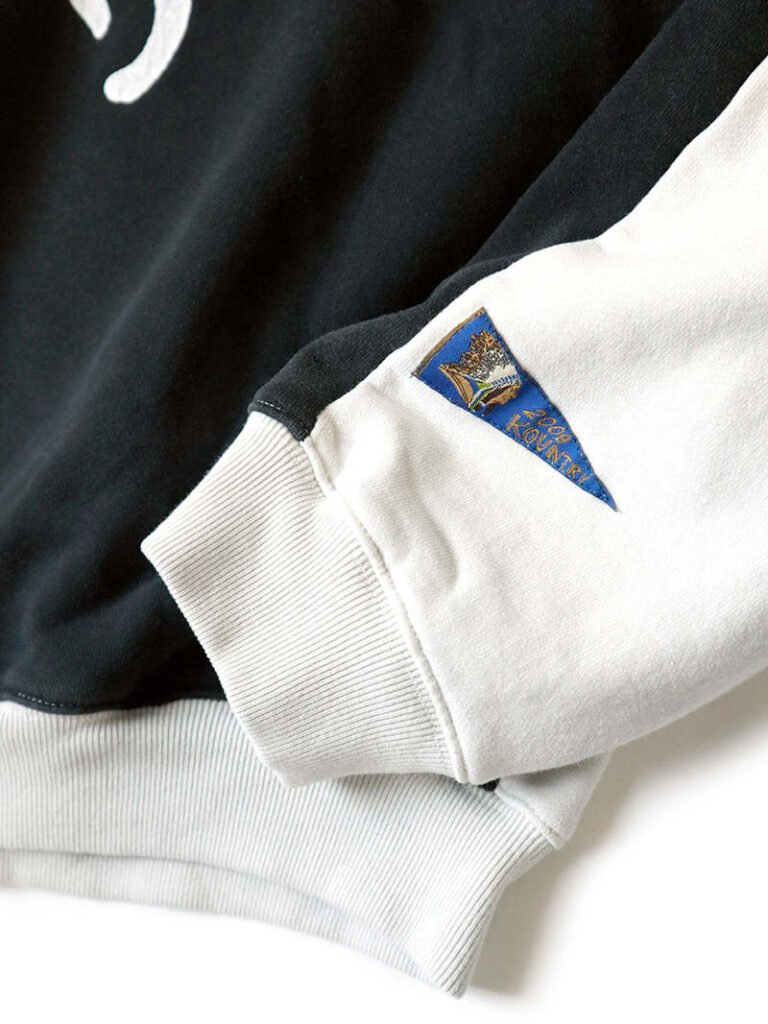 Close-up of the KAPITAL Men’s Sweat Knit 2Tones Remake Big Sweatshirt in Ecru and Black, highlighting the ribbed white cuff, contrasting black sleeve panel, and the signature blue KOUNTRY woven label stitched near the wrist.