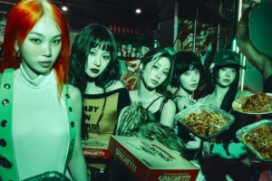 A stylized photo of five young women standing together in a dimly lit convenience store, illuminated by green neon light. They wear bold, futuristic streetwear with striking hairstyles — one with vivid orange hair — and hold boxes and trays of spaghetti. The scene blends cyberpunk aesthetics with playful, rebellious street culture energy.