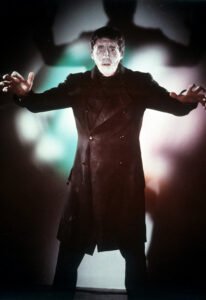 A dramatic color still of the Frankenstein Monster from Hammer Films’ The Curse of Frankenstein (1957). The creature, dressed in a dark, buttoned coat, extends his arms forward in a tense, almost pleading gesture. His pale, scarred face is illuminated by eerie green and red lighting, casting distorted shadows on the wall behind him that enhance the film’s gothic and unsettling atmosphere.
