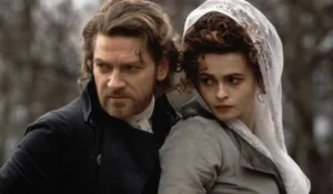 A still from the 1994 film Mary Shelley’s Frankenstein showing two characters in an outdoor setting. The man, dressed in dark 19th-century clothing with a stern expression, looks over his shoulder protectively, while the woman beside him, wearing a light gray dress and lace veil, gazes in the same direction with tension and concern. The muted tones and period costumes evoke the gothic atmosphere of the adaptation.