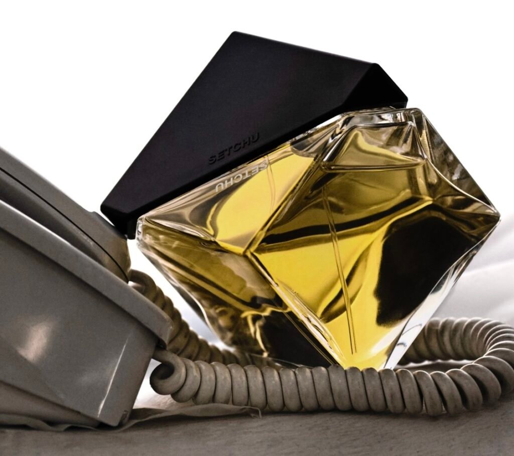 A sculptural glass perfume bottle filled with golden liquid, labeled “SETCHU,” featuring a sharp black geometric cap. The bottle rests against a coiled vintage telephone cord on a neutral surface, evoking a minimalist yet tactile sense of modern haute.
