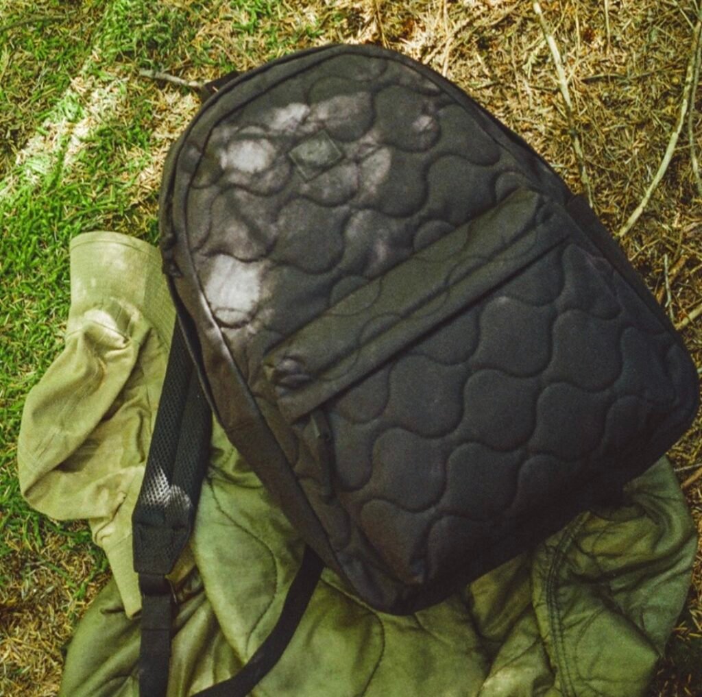 Close-up of a black Herschel Supply Co Quilted Capsule backpack resting on an olive quilted jacket in a grassy outdoor setting, highlighting the collection’s signature padded stitching and utilitarian, tactile design.