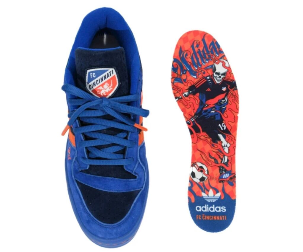 adidas Forum Low “All For Cincy” Blue Kit edition featuring the FC Cincinnati crest on the tongue, blue and orange suede upper with matching laces, and a custom insole illustrated with a fiery skeleton soccer player wearing the club’s kit, surrounded by smoke graphics and adidas FC Cincinnati branding at the heel.