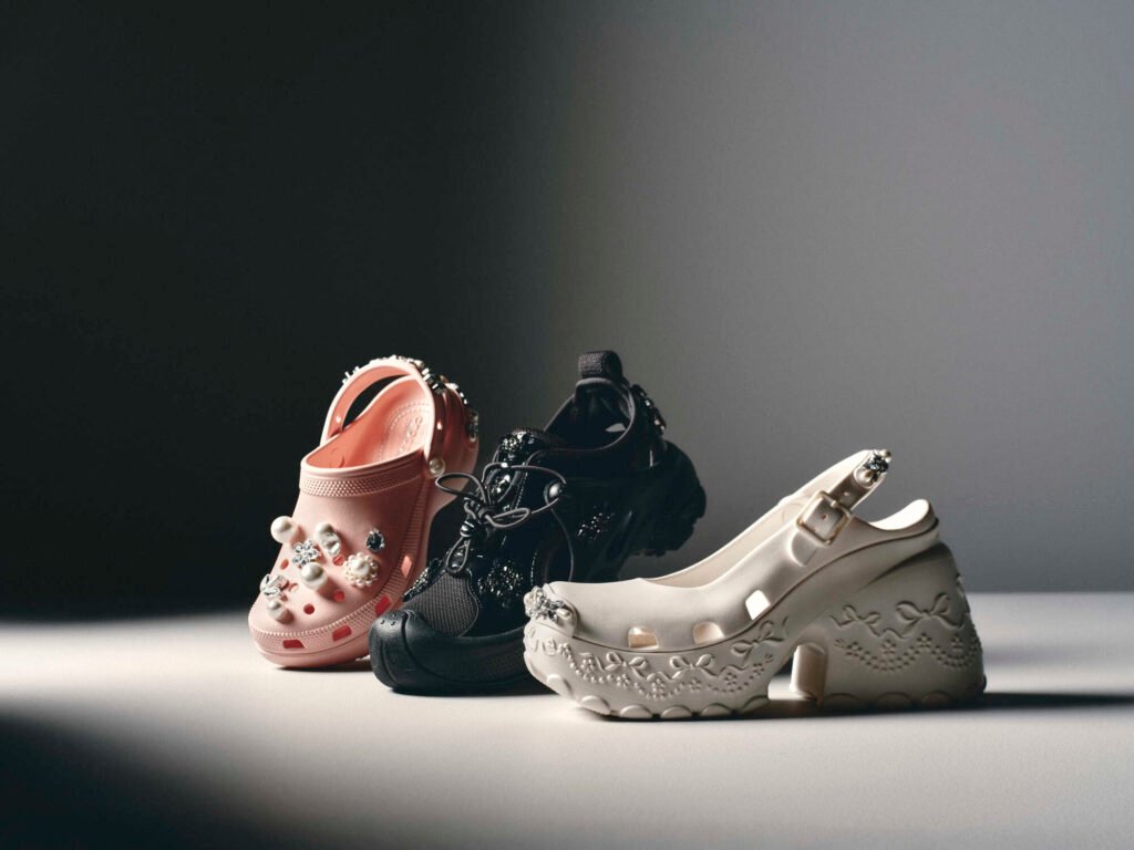Studio image showcasing the Simone Rocha x Crocs Spring/Summer 2026 collection. From left to right: the embellished Classic Platform Clog in Pink Rose adorned with pearls and crystals, the Trailbreak sneaker in Black featuring rugged lace detailing, and the new Ballerina Platform in Oyster with embossed bow motifs and a sculpted heel — blending Crocs’ utility with Simone Rocha’s romantic elegance.