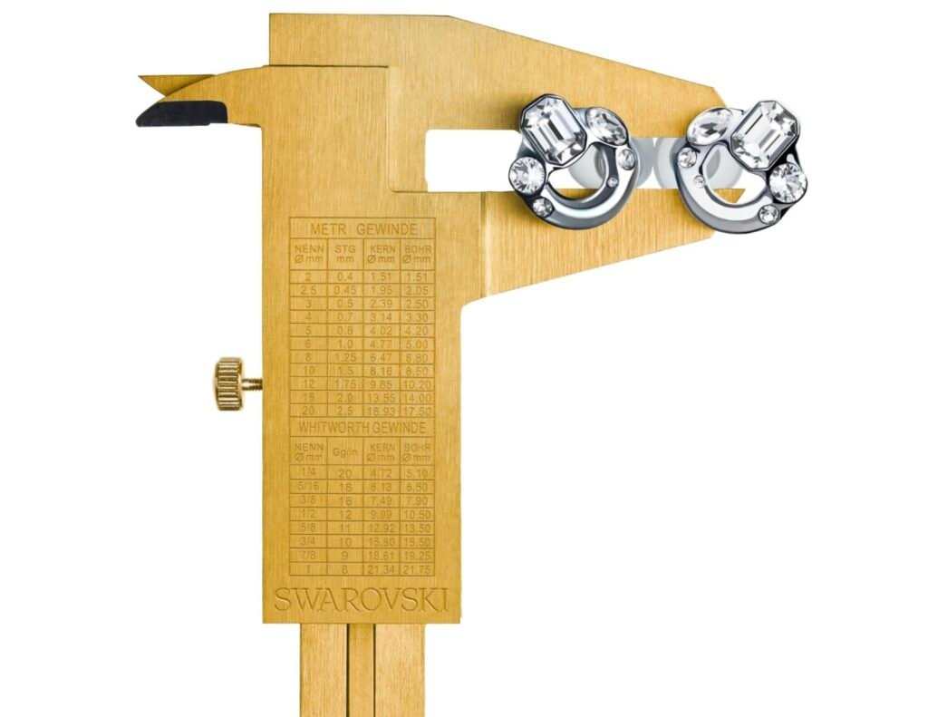 A gold caliper engraved with measurement markings and the Swarovski logo, used as a display stand holding a pair of Loop x Swarovski Experience 2 Limited Edition earplugs. The earplugs, encrusted with precision-cut Swarovski crystals, contrast against the matte gold instrument—symbolizing the collaboration’s blend of technical precision and luxurious craftsmanship.