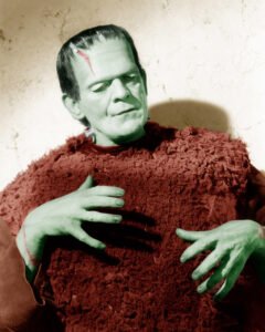 A colorized portrait of the Frankenstein Monster from Son of Frankenstein (1939). The creature, with pale green skin, heavy scars, and metal neck bolts, is shown in a textured reddish-brown garment. His hands are raised near his chest in a contemplative pose, and the soft lighting emphasizes both his iconic features and the tragic humanity beneath the monster’s expression.
