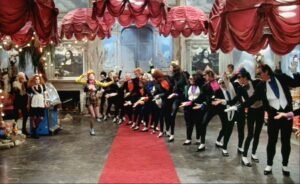 A colorful scene from The Rocky Horror Picture Show (1975) featuring a group of flamboyantly dressed characters performing a choreographed dance in a lavish hall. Red draped curtains hang from the ceiling, and a red carpet leads toward the center where a performer strikes a dramatic pose. The eclectic costumes, playful energy, and surreal décor reflect the film’s campy, theatrical, and rebellious spirit.