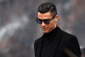 Cristiano Ronaldo wearing dark sunglasses and a black turtleneck with a blazer, looking down with a composed expression.