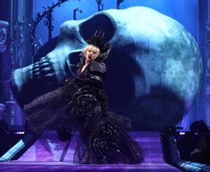 Lady Gaga performing at The MAYHEM Ball in London’s O2 Arena, dressed in a gothic black gown with oversized roses, singing in front of a giant illuminated skull backdrop.