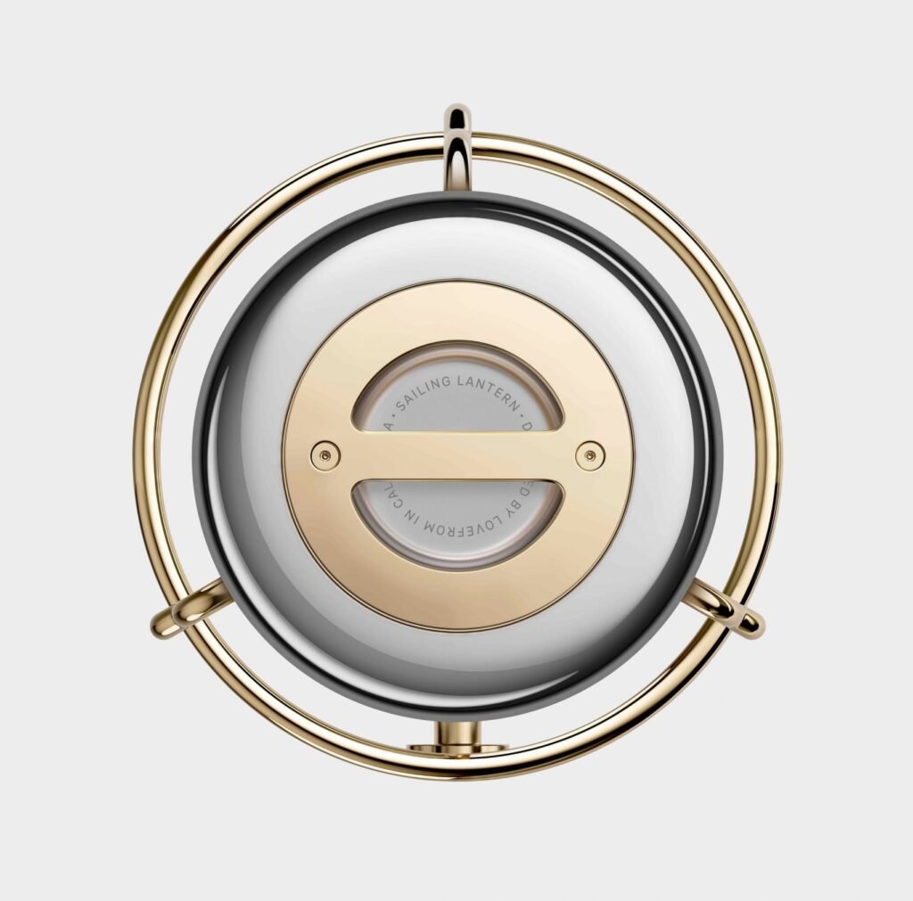 Top-down view of the Sailing Lantern designed by Jony Ive for Balmuda, featuring a polished circular metal body framed by a golden ring and minimalist detailing engraved with ‘Designed by LoveFrom in California.’ The composition highlights the lantern’s precision craftsmanship and nautical inspiration.