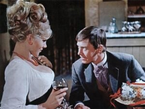 A scene from the 1957 film The Curse of Frankenstein showing a man and a woman in an intimate conversation at a dimly lit table. The man, dressed in a Victorian-style suit, gazes intently at the woman, who wears an elegant dress with pearls and holds a glass of wine. The warm lighting and rich colors highlight the film’s gothic yet sensual atmosphere, typical of Hammer Horror’s visual style.