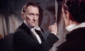 A color still from The Revenge of Frankenstein (1958) featuring a tense confrontation between two men in Victorian attire. The foreground figure, wearing a dark coat and crisp white shirt, stares coldly toward the other man while gripping a curtain or wall for emphasis. The dim lighting and sharp expressions underscore the film’s gothic tension and psychological intensity characteristic of Hammer Horror’s style.