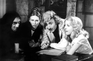 A black-and-white still from the 1974 comedy film Young Frankenstein, showing four characters gathered around a table in a dimly lit laboratory. The group, dressed in period costumes, leans in over handwritten notes, each with a distinct expression—ranging from suspicion to curiosity and amusement. The image captures the film’s blend of classic horror homage and satirical humor.