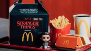 A McDonald’s Stranger Things meal displayed on a red tray, featuring a Hawkins-themed Happy Meal box with the Stranger Things logo, a small Eleven figurine, a box of fries, a burger container, and a McDonald’s drink cup against a warm red background