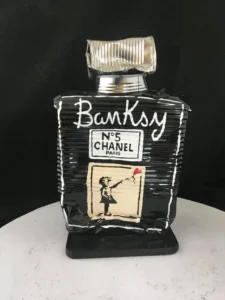 A black, crushed-metal sculpture shaped like a Chanel No. 5 perfume bottle, painted with the word “Banksy” in white and featuring a stencil-style image of a girl releasing a red heart-shaped balloon