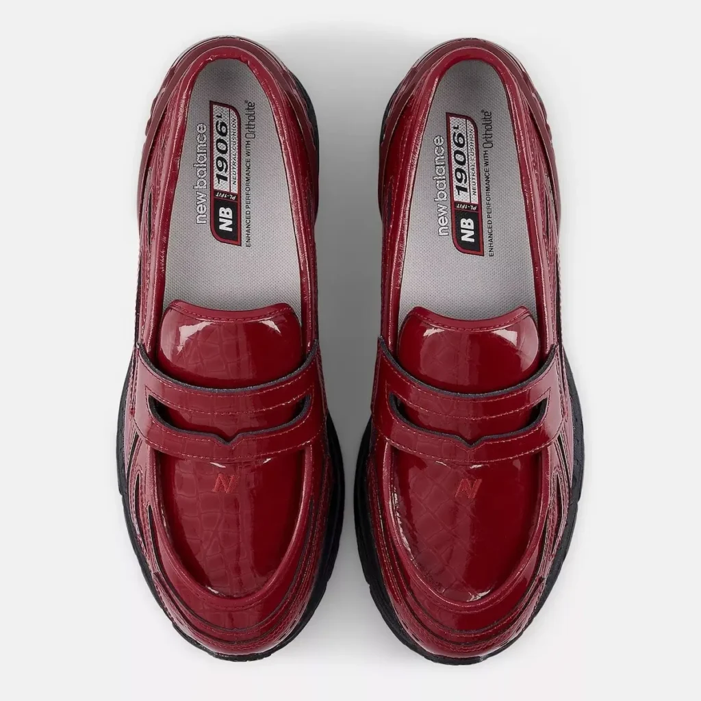 A top-down view of the New Balance 1906 LCS Loafer “Red Croc,” showcasing the glossy croc-embossed red patent upper, penny-strap saddle detail, and rounded apron-toe paneling. Both shoes are aligned symmetrically, revealing the grey OrthoLite insoles printed with “NB 1906” branding, while the sculpted black running-inspired midsole frames the silhouette from below