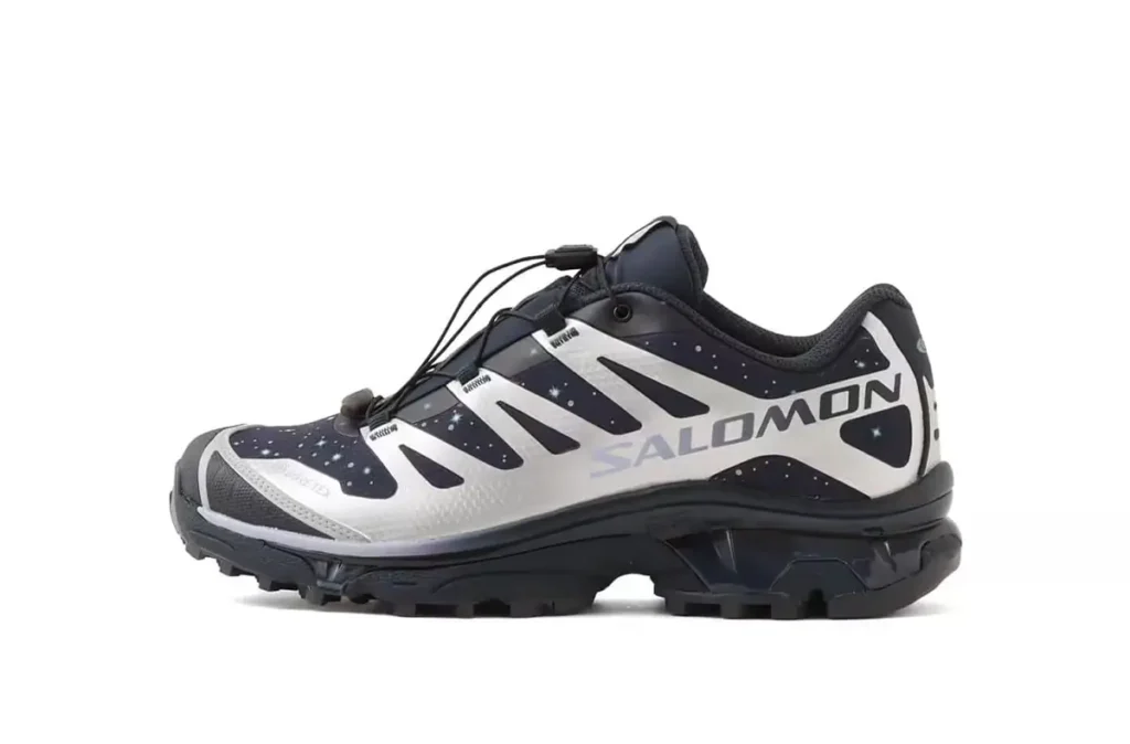 Side view of the atmos × Salomon XT-4 OG GTX “Stars Collide” sneaker showing its navy and black cosmic-speckled upper, silver TPU cage, bold SALOMON branding, and rugged black Contagrip trail outsole