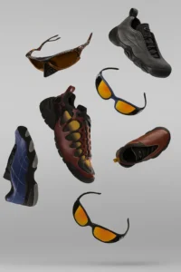 A floating arrangement of Oakley Factory Team FW25 footwear and eyewear displayed against a smooth grey backdrop. The composition features multiple pairs of sunglasses with amber-tinted lenses and sculptural frames, alongside several sneakers in red, grey, blue, and brown tones, each showcasing Oakley’s signature futuristic textures and paneling