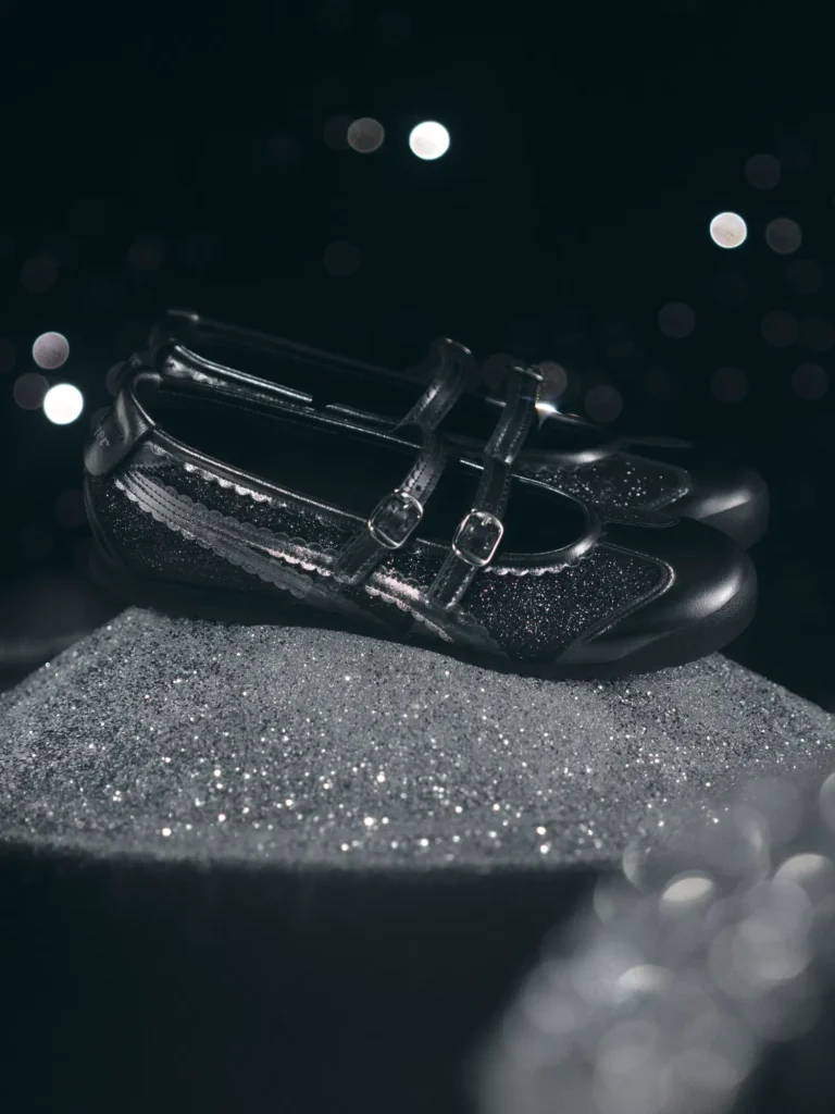A pair of black Onitsuka Tiger Mary Jane–style shoes with glittery panels sits on a textured, shimmering platform. The shoes feature double buckle straps and glossy leather edges, catching soft studio light. The dark background is dotted with blurred bokeh highlights, creating a dreamy, night-sky atmosphere around the sparkling footwear