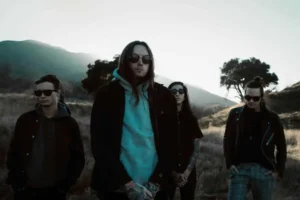 The band Bad Omens standing outdoors in a muted, cinematic landscape with mountains and dry grass behind them. All four members wear dark clothing and sunglasses, with the lead figure centered in a light gray hoodie and jacket. The evening light casts cool tones across the scene, giving the group a moody, atmospheric presence