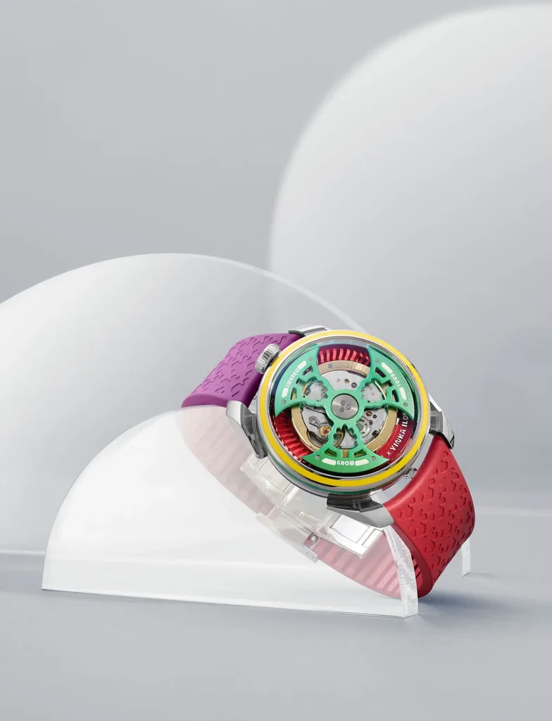 A Yinka Ilori × M.A.D. Editions × MB&F watch displayed on a clear half-dome stand, featuring a multicolored open-worked dial with green, yellow, and red accents. The timepiece is paired with a purple and red textured rubber strap embossed with Ilori’s signature pattern, set against a soft grey background with translucent curved forms