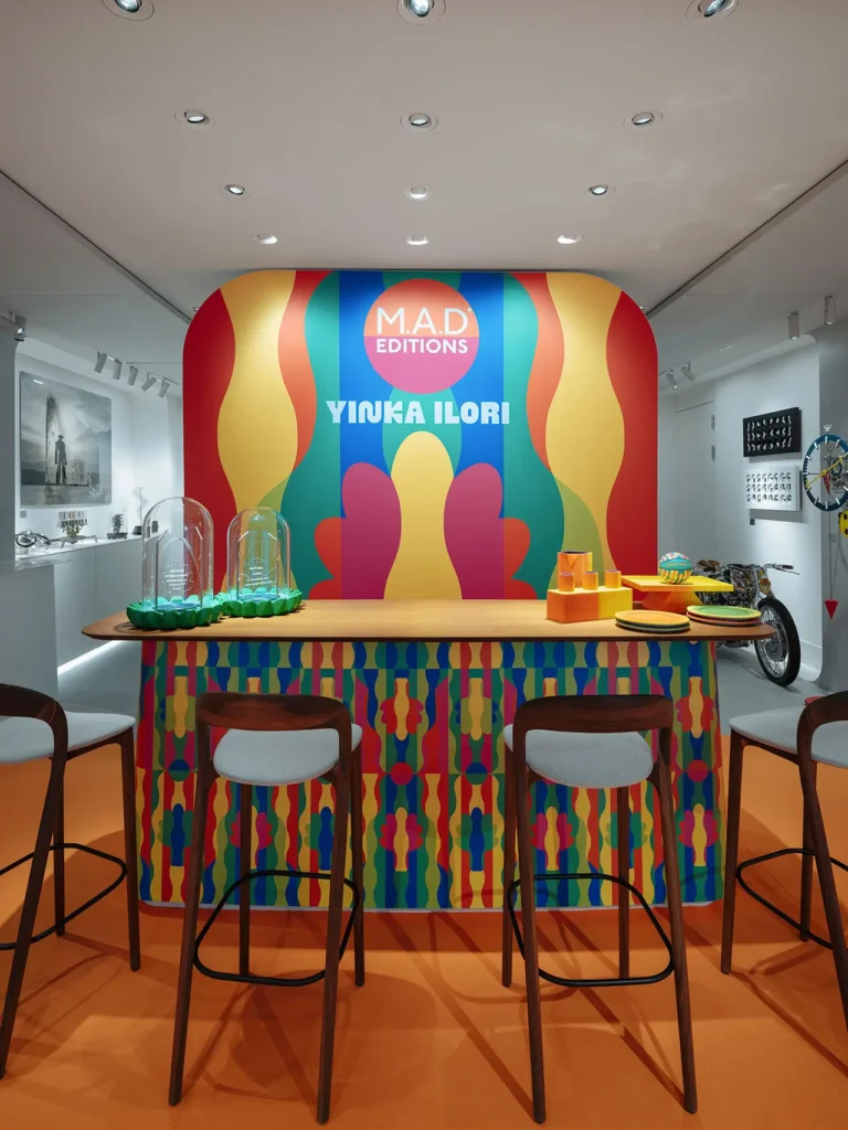 A brightly colored installation inside an MB&F M.A.D. Editions space, featuring a bar covered in Yinka Ilori’s signature geometric patterns in vibrant red, blue, yellow, and green. The backdrop displays the text “M.A.D. EDITIONS” and “YINKA ILORI” against a multicolored wavy design. Wooden bar stools with light grey cushions line the counter, while playful objects and domed display covers sit atop the bar, all set within a modern gallery-style environment