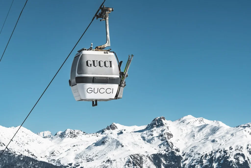 A white Gucci-branded ski gondola hangs above the snowy peaks of Courchevel under a clear blue sky, with skis mounted on its exterior